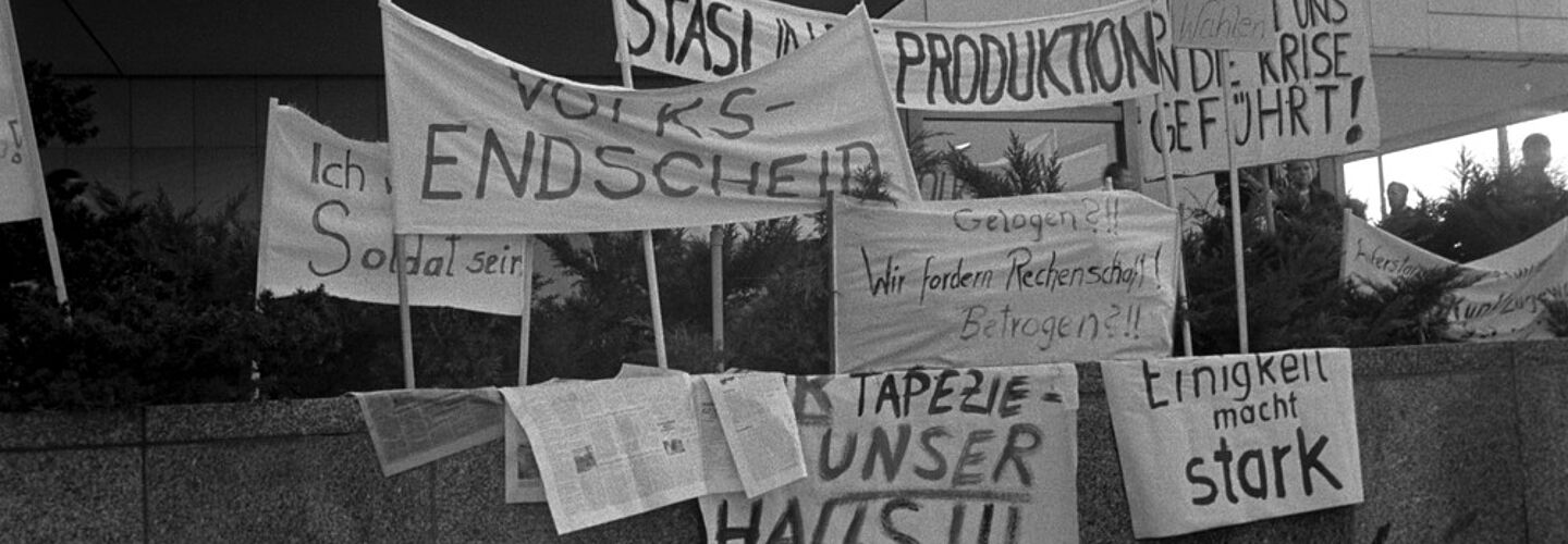 Auf einer Mauer sind mehrere Transparente aufgestellt. Die Losungen, soweit lesbar, lauten: 'Volksentscheid', 'Stasi in die Produktion', 'Ich will kein Soldat sein', 'Gelogen?!! Wir fordern Rechenschaft! Betrogen ?!!', 'Einigkeit macht stark' und 'Freie Wahlen'.